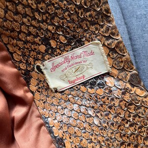May include: Close-up of a snakeskin pattern with brown and black scales. A tag reads "Specially Hand Made and fashioned by Ingeborg" in red script. The lining is a peach color, and a gray fabric is visible in the background.