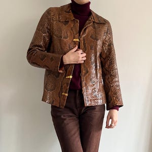 May include: A brown snakeskin-patterned jacket with a collared neckline and gold-colored clasps. The jacket is worn over a burgundy turtleneck sweater and paired with brown pants. The jacket's pattern features shades of brown and black.