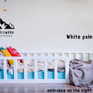 Peut inclure: Lit bébé en bois blanc avec un matelas bleu, des oreillers et une couverture grise. Le lit a un design à lattes et est placé contre un mur blanc. Le texte "HOLY-MOLYWOOD Family Carpentry Workshop" est sur le mur.