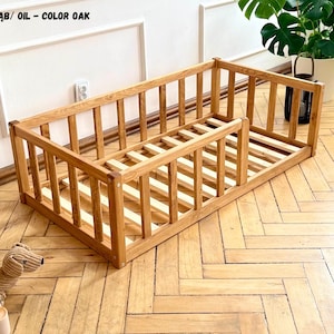 Op de afbeelding: Een rechthoekig houten vloerbedframe met een lattenbodem en een beschermend hekwerk aan drie zijden. Het frame heeft een eikenolie-afwerking. De tekst in de linkerbovenhoek luidt OLEJ - KOLOR DĄB/ OIL - COLOR OAK, rechtsonder staat een logo.