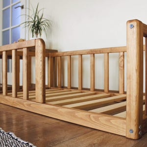 May include: A low-profile wooden floor bed frame featuring a slatted base and protective side railings. The natural wood grain is visible throughout the structure, which is assembled with exposed metal hardware at the corner joints.