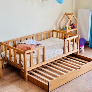 Könnte beinhalten: Ein Kinderbett aus Holz mit einer herausziehbaren Schublade, Sicherheitsgittern und einer gemusterten Matratze. Das Bett ist aus Naturholz gefertigt und hat ein schlichtes, modernes Design.