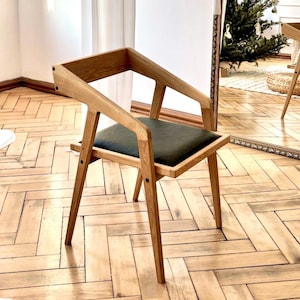 Op de afbeelding: Een houten stoel met een donkergroen zitkussen. De stoel heeft een uniek hoekig ontwerp met een lichtbruine afwerking. De afbeelding toont ook een grote spiegel, witte gordijnen en de tekst "HOLY-MOLYWOOD Family Carpentry Workshop".