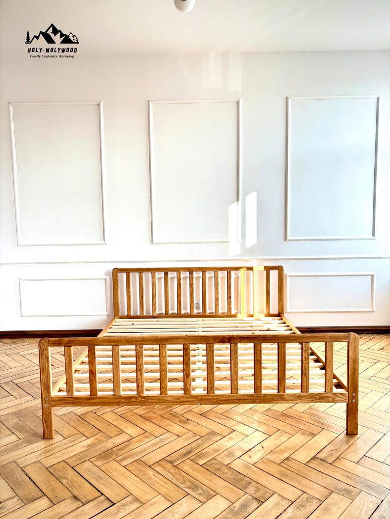 May include: A wooden bed frame with a slatted headboard and footboard, featuring a natural light brown finish and visible wood grain. The structure includes a slatted base for mattress support, with a logo in the top left corner reading HOLY-MOLYWOOD.