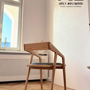 Op de afbeelding: Een houten stoel met een modern ontwerp. De stoel heeft een lichtbruine afwerking, een rechthoekige rugleuning en schuine armleuningen. De zitting lijkt bekleed te zijn met een donkergrijs materiaal. De tekst "HOLY-MOLYWOOD Family Carpentry Workshop" is zichtbaar.