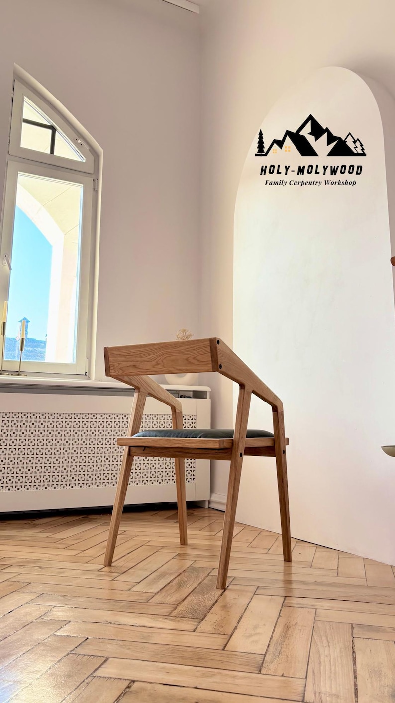 Op de afbeelding: Een houten stoel met een modern ontwerp. De stoel heeft een lichtbruine afwerking, een rechthoekige rugleuning en schuine armleuningen. De zitting lijkt bekleed te zijn met een donkergrijs materiaal. De tekst "HOLY-MOLYWOOD Family Carpentry Workshop" is zichtbaar.