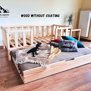 Könnte beinhalten: Ein hellfarbiger Holzbettenrahmen mit Kopf- und Seitenteilen. Das Bett besteht aus unbehandeltem Holz und hat eine Matratze mit Kissen und einer Decke. Ein schwarzes Dinosaurierspielzeug liegt auf der Decke. Der Text "WOOD WITHOUT COATING" steht über dem Bett.