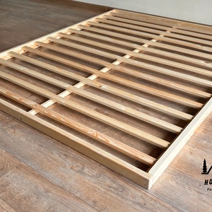 Op de afbeelding: Een houten bedframe met een lattenontwerp. Het frame is rechthoekig en gemaakt van lichtgekleurd hout. De latten zijn gelijkmatig verdeeld en lopen over de lengte van het frame. Het frame staat op een houten vloer.