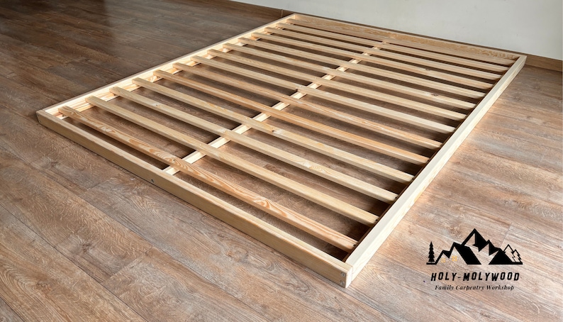 Op de afbeelding: Een houten bedframe met een lattenontwerp. Het frame is rechthoekig en gemaakt van lichtgekleurd hout. De latten zijn gelijkmatig verdeeld en lopen over de lengte van het frame. Het frame staat op een houten vloer.
