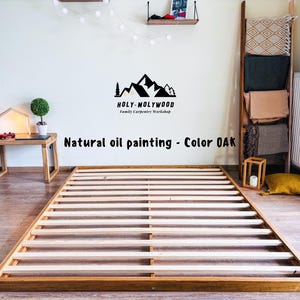 Op de afbeelding: Een houten bedframe met een lattenontwerp, gebeitst in een natuurlijke eikenkleur. Het frame is rechthoekig en staat op een lichtgekleurde vloer. De tekst "Natural oil painting - Color OAK" is zichtbaar.