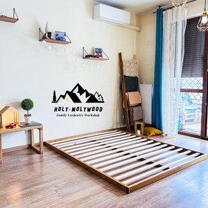 Op de afbeelding: Een houten bedframe met een natuurlijke eiken afwerking. Het frame is rechthoekig met een lattenbodem. De afbeelding toont ook een kleine bijzettafel, planken en de tekst "HOLY-MOLYWOOD Family Carpentry Workshop."