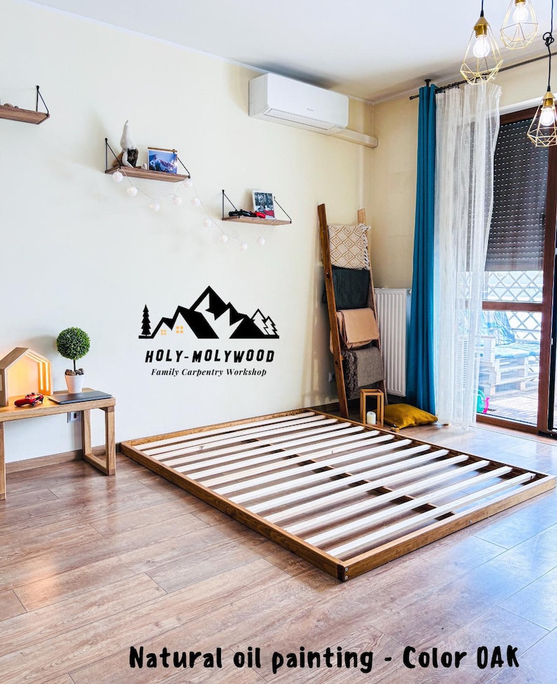 Op de afbeelding: Een houten bedframe met een natuurlijke eiken afwerking. Het frame is rechthoekig met een lattenbodem. De afbeelding toont ook een kleine bijzettafel, planken en de tekst "HOLY-MOLYWOOD Family Carpentry Workshop."