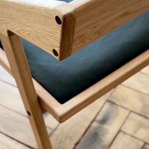 Op de afbeelding: Close-up van een houten stoel met een zwarte zitting. De stoel heeft een lichtbruine, natuurlijke houten afwerking en een modern design. Het frame van de stoel is geconstrueerd met zichtbare verbindingen en zwarte hardware. Het logo van Holy-Molywood Family Carpentry Workshop is zichtbaar.