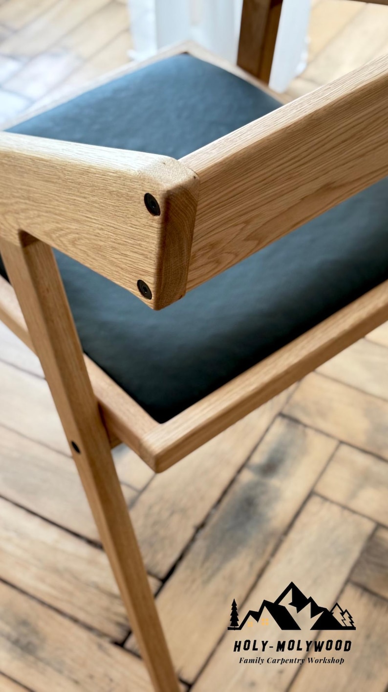 Op de afbeelding: Close-up van een houten stoel met een zwarte zitting. De stoel heeft een lichtbruine, natuurlijke houten afwerking en een modern design. Het frame van de stoel is geconstrueerd met zichtbare verbindingen en zwarte hardware. Het logo van Holy-Molywood Family Carpentry Workshop is zichtbaar.