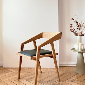 Op de afbeelding: Houten fauteuil met een donkergrijze zitting. De stoel heeft een modern design met schuine armleuningen en poten. De achtergrond toont een witte muur met de tekst "HOLY-MOLYWOOD Family Carpentry Workshop".