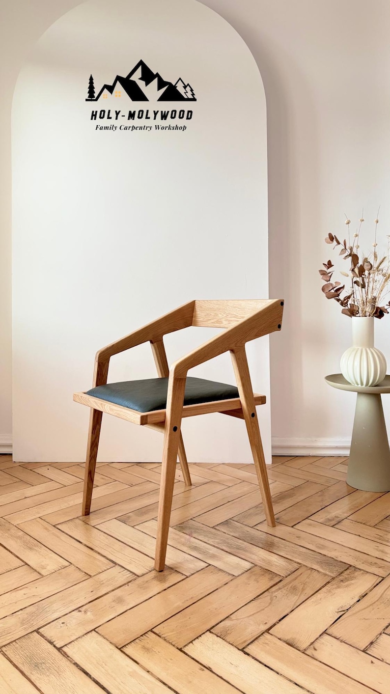 Op de afbeelding: Houten fauteuil met een donkergrijze zitting. De stoel heeft een modern design met schuine armleuningen en poten. De achtergrond toont een witte muur met de tekst "HOLY-MOLYWOOD Family Carpentry Workshop".