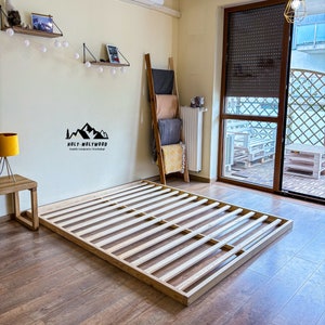 Op de afbeelding: Een houten bedframe met een lattenbodem ontwerp, rustend op een houten vloer. Het frame is rechthoekig en lijkt van lichtgekleurd hout te zijn. Het Holy-Molywood Family Carpentry Workshop logo is zichtbaar.