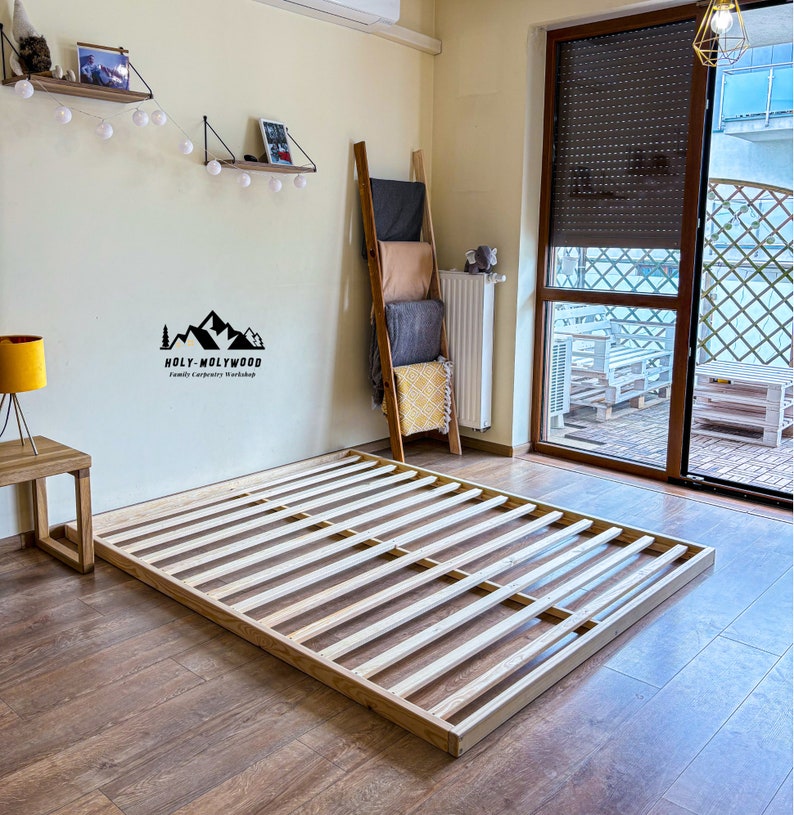 Op de afbeelding: Een houten bedframe met een lattenbodem ontwerp, rustend op een houten vloer. Het frame is rechthoekig en lijkt van lichtgekleurd hout te zijn. Het Holy-Molywood Family Carpentry Workshop logo is zichtbaar.
