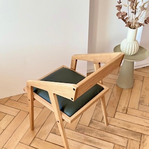 Op de afbeelding: Houten stoel met een donkergroen zitkussen. De stoel heeft een modern ontwerp met schuine poten en rugleuningen. Het logo "HOLY-MOLYWOOD Family Carpentry Workshop" is zichtbaar. De stoel staat op een houten vloer.