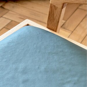 Op de afbeelding: Close-up van een houten stoel met een lichtblauwe leren zitting. De stoel heeft een natuurlijke houten frame en een vierkante zitting. De afbeelding bevat de tekst "HOLY-MOLYWOOD Family Carpentry Workshop".