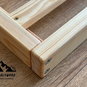 Op de afbeelding: Close-up van een houten framehoek, met de natuurlijke nerf van het lichtgekleurde hout. Het frame is met schroeven verbonden en lijkt deel uit te maken van een grotere structuur. Het logo "HOLY-MOLYWOOD Family Carpentry Workshop" is zichtbaar.