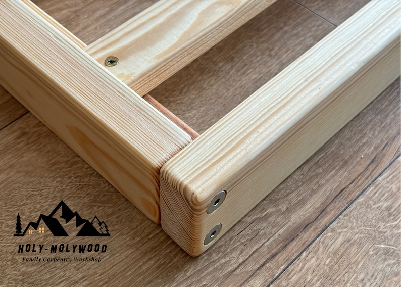 Op de afbeelding: Close-up van een houten framehoek, met de natuurlijke nerf van het lichtgekleurde hout. Het frame is met schroeven verbonden en lijkt deel uit te maken van een grotere structuur. Het logo "HOLY-MOLYWOOD Family Carpentry Workshop" is zichtbaar.