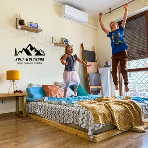 Op de afbeelding: Een laag houten bedframe met een matras, kussens en een patroon deken. Twee kinderen springen op het bed. Een houten bijzettafel met een lamp en een logo "HOLY-MOLYWOOD Family Carpentry Workshop" zijn ook zichtbaar.