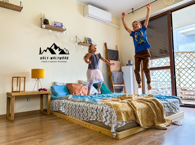 Op de afbeelding: Een laag houten bedframe met een matras, kussens en een patroon deken. Twee kinderen springen op het bed. Een houten bijzettafel met een lamp en een logo "HOLY-MOLYWOOD Family Carpentry Workshop" zijn ook zichtbaar.