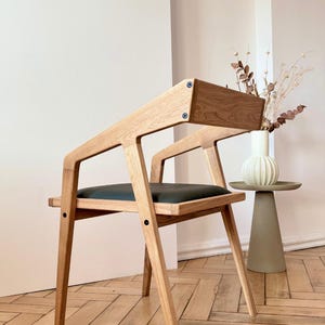 Op de afbeelding: Een moderne houten stoel met een donkergroene zitting en een licht houten frame. De stoel heeft een uniek schuin ontwerp en staat tegen een witte muur. De tekst "HOLY-MOLYWOOD Family Carpentry Workshop" is zichtbaar.