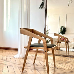 Op de afbeelding: Een houten stoel met een modern ontwerp. De stoel heeft een lichtbruin frame, een donkergrijze zitting en schuine armleuningen. De stoel staat op een houten vloer. Het logo "HOLY-MOLYWOOD Family Carpentry Workshop" is zichtbaar.