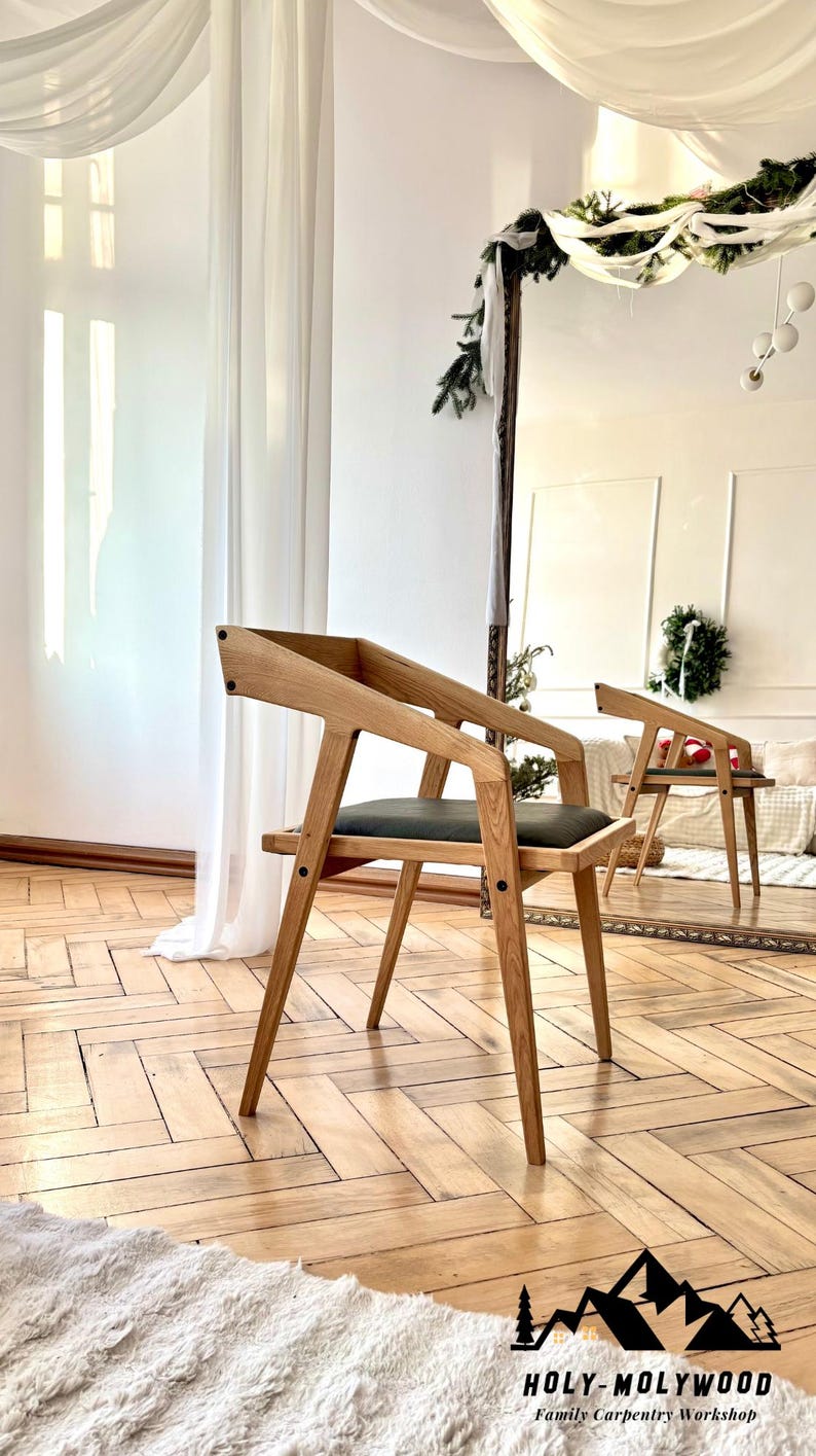 Op de afbeelding: Een houten stoel met een modern ontwerp. De stoel heeft een lichtbruin frame, een donkergrijze zitting en schuine armleuningen. De stoel staat op een houten vloer. Het logo "HOLY-MOLYWOOD Family Carpentry Workshop" is zichtbaar.