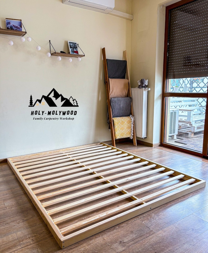 Op de afbeelding: Een houten bedframe met een lattenbodem ontwerp. Het frame is rechthoekig en staat op een houten vloer. De muur achter het bedframe heeft een logo met de tekst "HOLY-MOLYWOOD Family Carpentry Workshop".