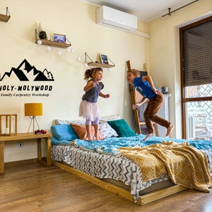 Op de afbeelding: Een houten bedframe met een matras, kussens en dekens. Twee kinderen springen op het bed. De muur heeft een logo met de tekst "HOLY-MOLYWOOD Family Carpentry Workshop". Een houten bijzettafel met een lamp is ook zichtbaar.
