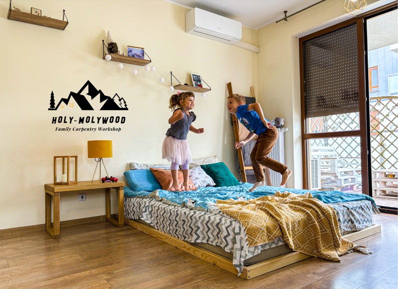 Op de afbeelding: Een houten bedframe met een matras, kussens en dekens. Twee kinderen springen op het bed. De muur heeft een logo met de tekst "HOLY-MOLYWOOD Family Carpentry Workshop". Een houten bijzettafel met een lamp is ook zichtbaar.