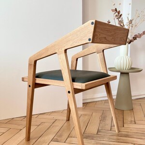 Op de afbeelding: Houten stoel met een donkergroene zitting en een unieke schuine rugleuning. De stoel is gemaakt van lichtgekleurd hout en heeft een modern design. De tekst "HOLY-MOLYWOOD Family Carpentry Workshop" is zichtbaar.