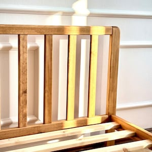 May include: A light brown wooden bed frame featuring a headboard with vertical slats and a slatted base. The headboard has a smooth, rounded top edge. The logo HOLY-MOLYWOOD Family Carpentry Workshop is visible in the upper left corner of the image.