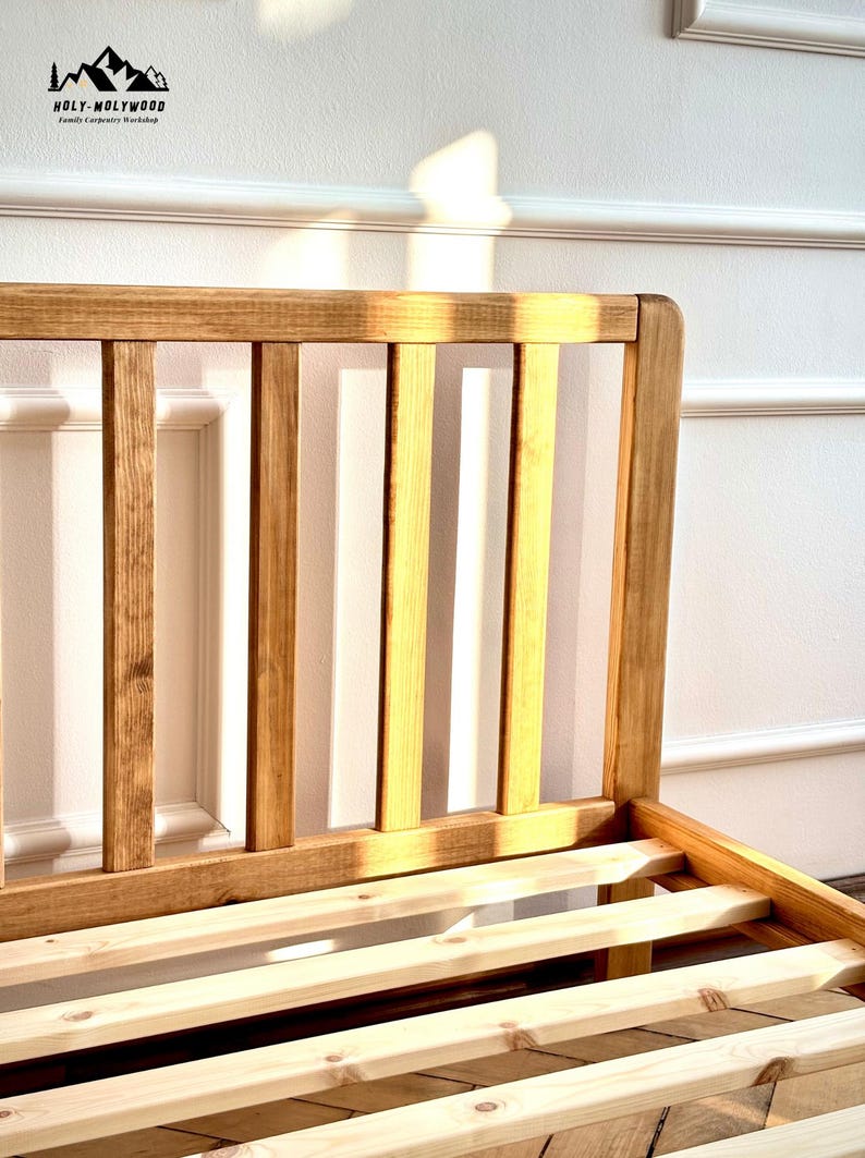 May include: A light brown wooden bed frame featuring a headboard with vertical slats and a slatted base. The headboard has a smooth, rounded top edge. The logo HOLY-MOLYWOOD Family Carpentry Workshop is visible in the upper left corner of the image.