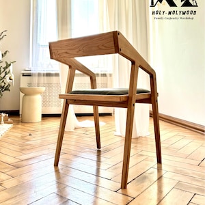 Op de afbeelding: Houten stoel met een minimalistisch ontwerp. De stoel heeft een lichtbruin houten frame en een donkergroen zitkussen. De rugleuning en armleuningen zijn in &eacute;&eacute;n stuk ge&iuml;ntegreerd. De stoel staat op een houten vloer.