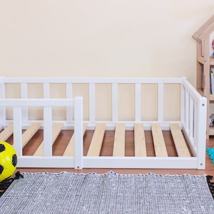 May include: White wooden toddler bed frame with a slatted base and a removable side rail. The bed is in a room with a toy shelf and a rug on the floor.