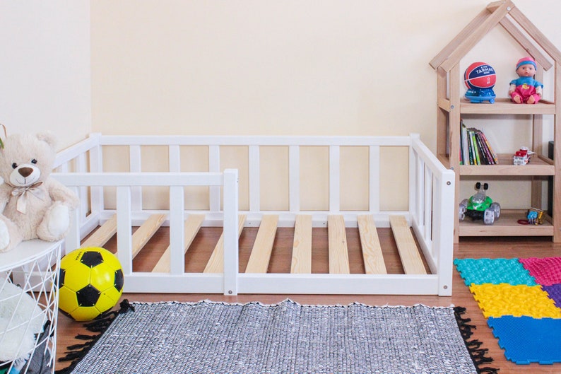 May include: White wooden toddler bed frame with a slatted base and a removable side rail. The bed is in a room with a toy shelf and a rug on the floor.