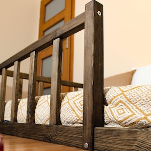 May include: A dark brown wooden dog bed with a white and yellow patterned blanket. The bed has a raised wooden frame with vertical slats.