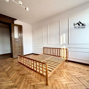 May include: A light brown wooden bed frame with a slatted headboard and footboard, featuring a natural oak oil finish. The frame is constructed with vertical wooden slats and sits on a herringbone patterned wooden floor in a room with white walls.