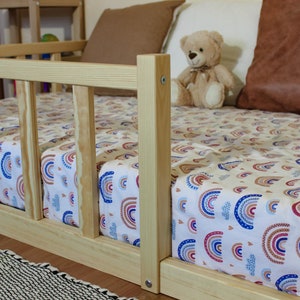 May include: A wooden toddler bed with a rainbow patterned sheet and a teddy bear sitting on the bed.