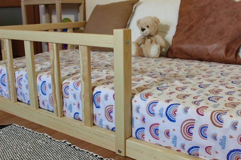 May include: A wooden toddler bed with a rainbow patterned sheet and a teddy bear sitting on the bed.