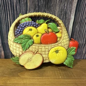 May include: A decorative wall hanging featuring a woven basket overflowing with colorful fruit. The basket contains grapes, pears, apples, and leaves. A sliced apple and a whole apple are placed in front of the basket. The colors are vibrant and the style is vintage.