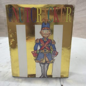 Peut inclure: Une boîte rectangulaire dorée avec le texte THE NUTCRACKER en lettres multicolores en haut. Le centre présente une illustration dessinée à la main d'un soldat en uniforme bleu avec des sangles croisées rouges, un grand chapeau et une épée.