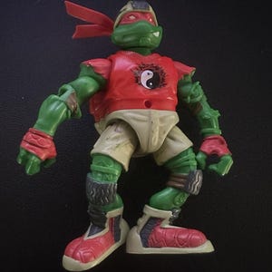 May include: A Teenage Mutant Ninja Turtle action figure. The figure is green with a red mask, red wrist and ankle guards, and red and white shoes. It wears a red shirt with a yin-yang symbol and khaki shorts.