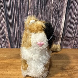 May include: A plush toy cat with a calico pattern of brown, white, and black fur. The cat has a pink nose and a small tail. The toy is sitting on a wooden surface.