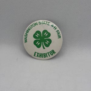 May include: A round white pinback button featuring a green four-leaf clover emblem with the letter H in each leaf. The text WASHINGTON STATE 4-H FAIR is printed in green along the top edge, and the word EXHIBITOR is printed in green at the bottom.