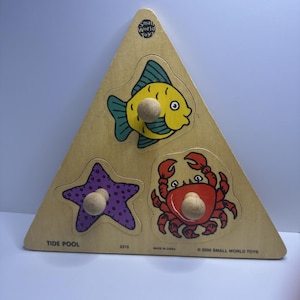 May include: Wooden triangular puzzle featuring a yellow fish, red crab, and purple starfish. Each piece has a wooden knob for easy grasping. The puzzle is labeled "Tide Pool" and includes the text "© 2000 SMALL WORLD TOYS."